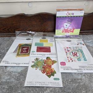 May include: A selection of Sizzix die-cutting sets, featuring floral, card, and seasonal designs. The sets include various shapes and sizes, with some featuring the text "YOU & ME" and "LOVE". The packaging is white with colourful illustrations.