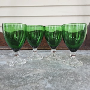 May include: Four green-tinted wine glasses with clear, textured bases and stems. The glasses are arranged in a row, showcasing their symmetrical design. The green color is consistent throughout the glass, creating a cohesive look. The glasses are likely vintage.