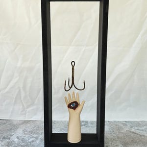May include: A tall, rectangular black wooden frame containing a small, pale mannequin hand with a painted blue eye in the center of the palm. A large, rusted metal triple-pronged fishing hook hangs suspended from the top of the frame above the hand.