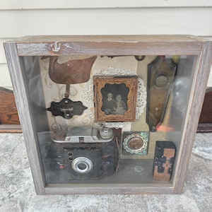 May include: A shadow box with a wooden frame, showcasing vintage items. Inside, a black camera, a small framed photograph, and decorative metal pieces are arranged against a patterned backdrop. The box has a glass front.