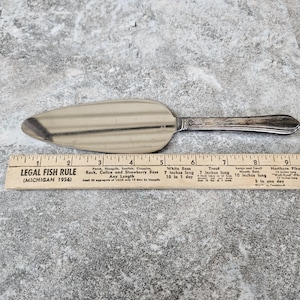 May include: A silver pie server with a decorative handle. The server is placed next to a vintage ruler with text about fish sizes and regulations. The ruler is marked with inches.