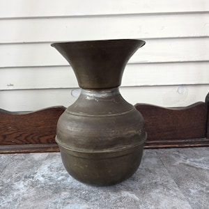 Puede incluir: Jarrón antiguo de latón con base bulbosa y parte superior acampanada. El jarrón tiene un aspecto deslustrado y envejecido, con desgaste y pátina visibles. El diseño presenta bandas horizontales alrededor del cuerpo. Una pieza decorativa.