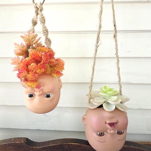 May include: Two hanging planters made from doll heads, each suspended by a woven rope. One planter holds orange and yellow flowers, the other holds a green succulent. The doll heads are painted with realistic features.
