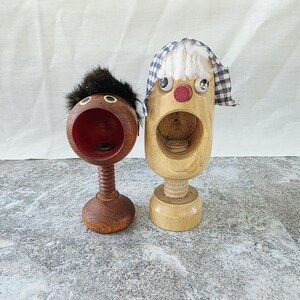 May include: Two wooden nutcrackers with spring-loaded mechanisms. The left nutcracker is dark brown with black hair and a red interior. The right nutcracker is light wood with white yarn hair, a blue and white checkered head covering, and a red nose.
