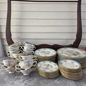Puede incluir: Un juego de té vintage con diseños florales. El juego incluye tazas de té, platillos y platos con un borde azul y ribete dorado. La disposición está sobre una superficie de madera con un estante decorativo de madera en el fondo.