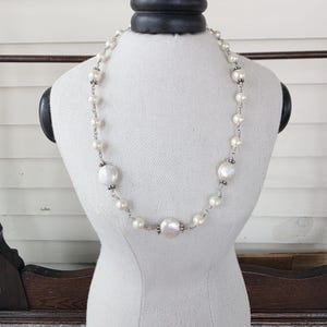 May include: A pearl necklace displayed on a white mannequin. The necklace features a mix of small and large, irregularly shaped white pearls strung together with a silver chain. The pearls vary in size, creating a unique and elegant design.