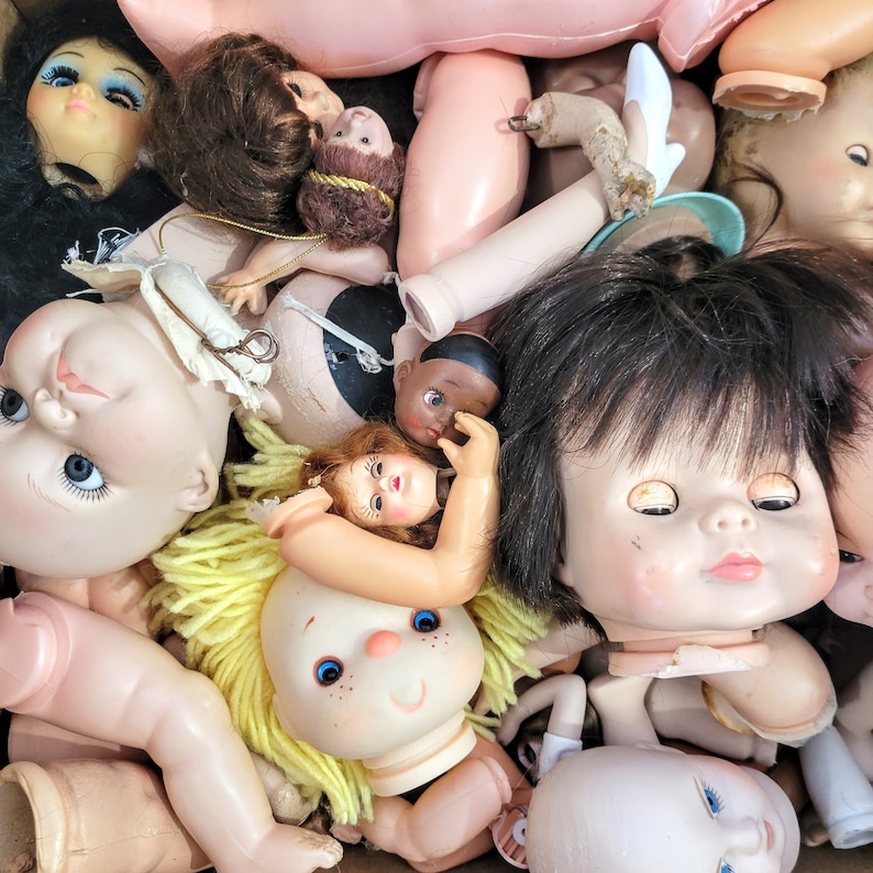 May include: A collection of vintage porcelain dolls with various hairstyles and clothing. The dolls are in various poses and have painted facial features. Some dolls have blonde hair, while others have brown hair. The dolls are in a variety of colors, including pink, white, and brown.