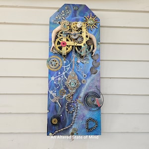 May include: A blue and gold mixed media artwork featuring a clock face with gears and a doll's head. The artwork is titled "An Altered State of Mind".
