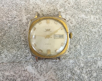 Op de afbeelding: Vintage Westclox polshorloge met een goudkleurige vierkante kast en een witte wijzerplaat. Het horloge heeft gouden wijzers en markeringen, een dag-datumweergave en de tekst "17 JEWELS". Het horloge staat op donderdag de 13e.