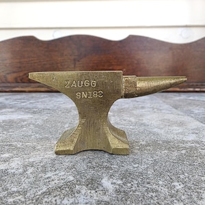 May include: A small, gold-colored metal anvil with the text "ZAUGG SN183" stamped on the front. The anvil has a flat top, a pointed horn, and a wide base. The background is a weathered wooden surface.