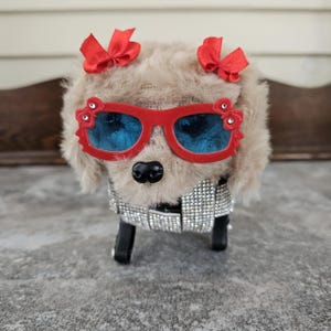 May include: A plush dog figure with a light brown fur coat, red sunglasses, and two red bows. The dog is wearing a silver rhinestone-studded jacket. The sunglasses have blue lenses.