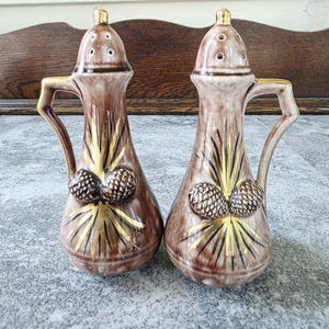 May include: A pair of ceramic salt and pepper shakers featuring a brown glazed finish with gold-painted pinecone and needle motifs. Each shaker has a handle, a tapered body, and a gold-toned lid with small holes for dispensing seasoning.