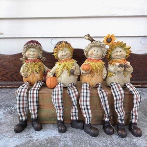 May include: Four whimsical fall figurines with smiling faces, leaf-like collars, and plaid-patterned legs. Each holds a pumpkin or sunflower. The figurines are seated on a wooden bench, perfect for autumn decor.