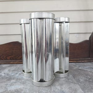 May include: Three vintage, silver-colored metal tube molds with fluted sides. The molds have a cylindrical shape with a flat base and a lid. They are arranged on a gray surface, with a wooden background.