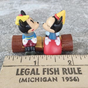 Może przedstawiać: Figurki Myszki Miki i Myszki Minnie, ubrane w niebieskie i czerwone stroje, zwr&oacute;cone twarzami do siebie na brązowej ławce z kłody. Figurki są czarne, białe i czerwone. Linijka z napisem "LEGAL FISH RULE (MICHIGAN 1956)" znajduje się na pierwszym planie.