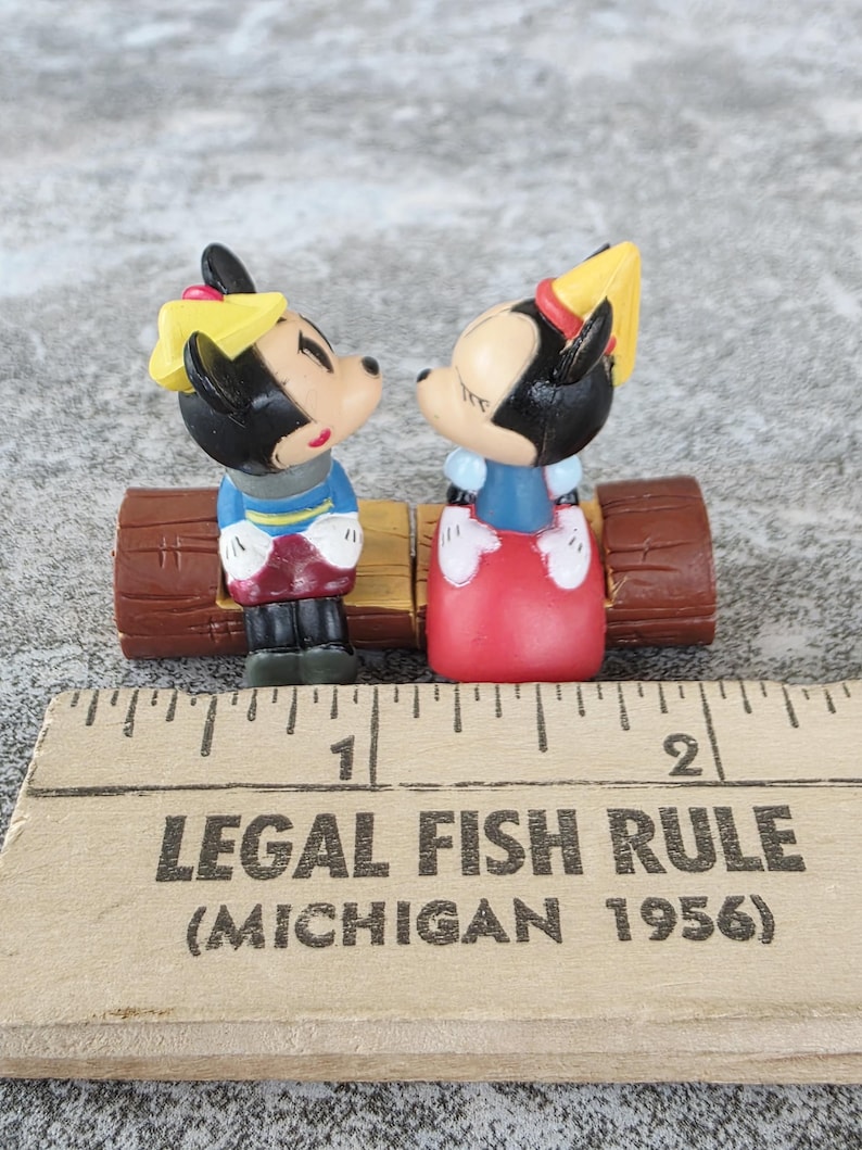 Może przedstawiać: Figurki Myszki Miki i Myszki Minnie, ubrane w niebieskie i czerwone stroje, zwr&oacute;cone twarzami do siebie na brązowej ławce z kłody. Figurki są czarne, białe i czerwone. Linijka z napisem "LEGAL FISH RULE (MICHIGAN 1956)" znajduje się na pierwszym planie.