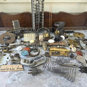 May include: A collection of assorted vintage hardware, metal components, and mechanical parts including hinges, springs, a brass pipe, a wooden ruler marked 6 inches, and various fasteners scattered on a surface in front of a wooden frame.