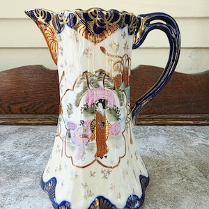 May include: A ceramic jug with a cream-coloured body, featuring a scalloped rim and base trimmed in deep blue and gold. The surface is decorated with gold star accents and a central illustration of two figures in traditional attire under a pink umbrella.
