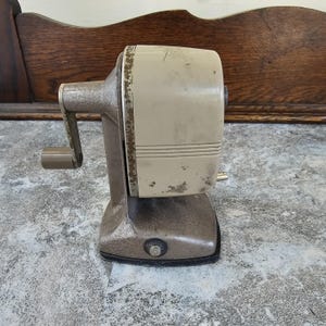 May include: A vintage pencil sharpener with a cream-colored body and a metallic gray base and handle. The sharpener shows signs of wear, with some paint chipping. The design is a classic, functional piece.