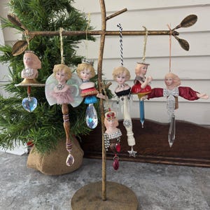 Puede incluir: Una colección de adornos navideños de estilo vintage, cada uno con una cara de muñeca y elaborados adornos. Los adornos están suspendidos de un árbol decorativo de color dorado, con una variedad de colores y texturas, incluyendo cristales, tela y cuentas.