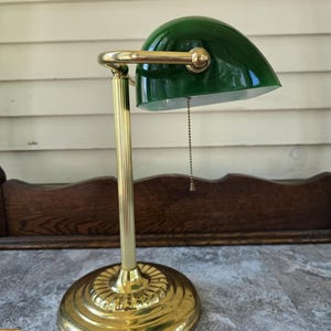 May include: A banker's lamp featuring a polished brass base and a vertical fluted stem. The lamp has a semi-cylindrical green glass shade with a white interior and a gold-toned metal pull chain hanging from the center for operation.