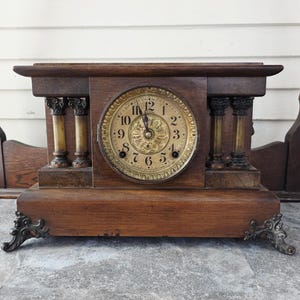 May include: Antique wooden mantel clock with a round clock face. The clock has a dark brown wooden frame with decorative columns and ornate metal feet. The clock face features Roman numerals and decorative hands.