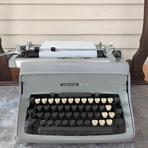 May include: A vintage Underwood Five typewriter in light gray, with black and cream-colored keys. A sheet of paper is inserted, ready for typing. The typewriter sits on a textured surface.