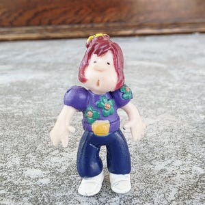 May include: A small plastic figurine of a person with red hair and a purple top with green flower accents. The figurine wears dark blue trousers, a yellow belt, and white shoes. The figure has a neutral expression.