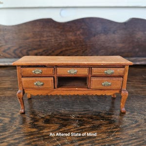 Puede incluir: Una cómoda de madera en miniatura con seis cajones y un estante. La cómoda tiene un tinte marrón y tiradores de cajones de latón. La cómoda está sobre una superficie de madera.