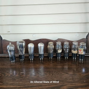May include: A row of ten vintage glass vacuum tubes with metal bases, arranged on a wooden surface. The tubes are clear glass with a silver coating on the inside. Some of the tubes have labels with text.