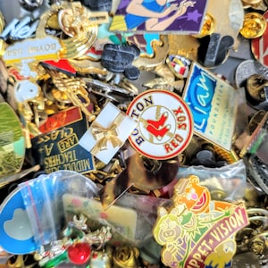May include: A collection of colorful enamel pins featuring various designs, including a baseball pin with the text "BO TON JOX", a pin with the text "Liam FOUNDATION", and a pin with a cartoon character.