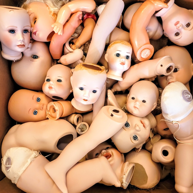 May include: A collection of porcelain doll heads and body parts in various poses. The heads are painted with realistic features and have different hairstyles. Some of the body parts are painted in a flesh tone, while others are white.