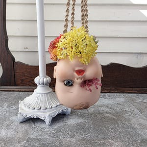 May include: A unique hanging planter made from a doll's head, adorned with yellow and red flowers. The doll head is suspended by macrame rope, creating a whimsical and artistic display. The planter is a creative home decor item.