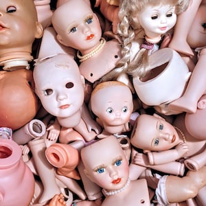 May include: A collection of porcelain dolls with various facial expressions and hairstyles. The dolls are arranged in a pile, with some of them partially obscured by others. The dolls are all wearing different outfits, including dresses, skirts, and pants.