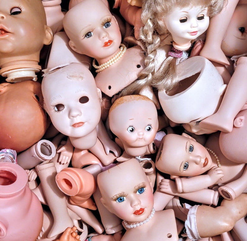 May include: A collection of porcelain dolls with various facial expressions and hairstyles. The dolls are arranged in a pile, with some of them partially obscured by others. The dolls are all wearing different outfits, including dresses, skirts, and pants.