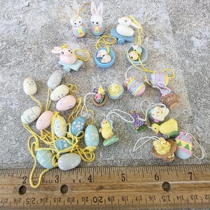 May include: A selection of Easter decorations featuring bunnies, chicks, and painted eggs in pastel shades. Each ornament has a yellow string for hanging. A wooden ruler with inch measurements is in the foreground.