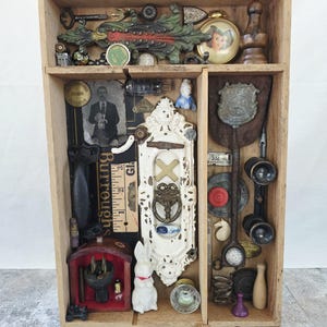 May include: A wooden shadow box display case filled with an assortment of vintage objects including a white ornate door plate, a black and white photograph of a man, a small white rabbit figurine, metal keys, a badge, and various hardware pieces.