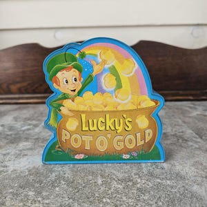 May include: A colourful, die-cut sign featuring a cartoon leprechaun reaching towards a rainbow. The sign reads "Lucky's POT O' GOLD" and depicts a pot overflowing with gold coins. The background is blue.