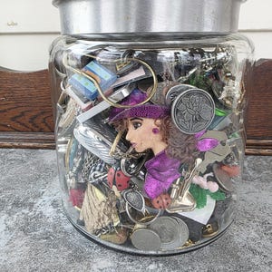 Puede incluir: Un tarro de cristal transparente con tapa plateada lleno de varios objetos pequeños. En el interior hay joyas, monedas y objetos decorativos. Se ve una figura de muñeca con sombrero y bufanda morados. El tarro está sobre una superficie gris.