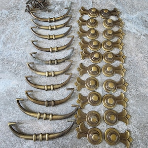 May include: A collection of antique brass-toned metal hardware including nine curved drawer pulls with central detailing and ten matching circular knobs with decorative leaf-shaped backplates, accompanied by a small pile of metal mounting screws.