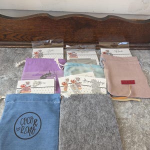 May include: An assortment of small drawstring bags in various colors, including blue, purple, and gray, with attached tags. Some bags feature the text "Coco & Reno" and a heart logo. The tags display decorative elements and handwritten script.
