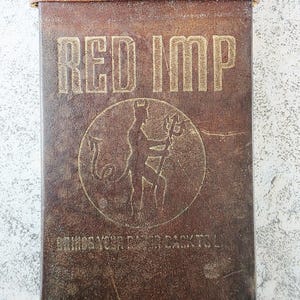 Puede incluir: Una funda de cuero marrón con las palabras "RED IMP" en letras doradas. Debajo, un emblema circular con una figura de diablo sosteniendo un tridente. La parte superior de la funda es de cuero marrón claro.