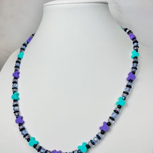 May include: A beaded necklace featuring purple, teal, and black beads. The necklace has a repeating pattern of purple bear-shaped beads and teal beads, interspersed with smaller black and iridescent beads. The necklace is displayed on a white mannequin.