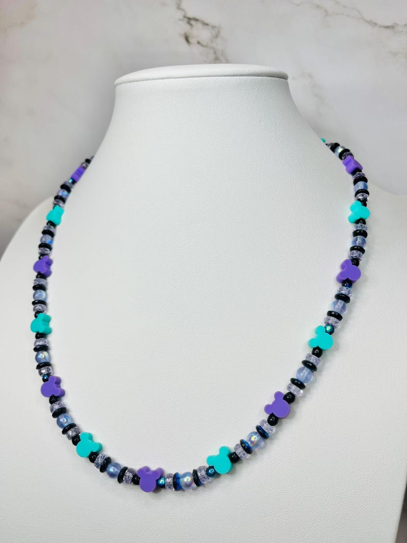 May include: A beaded necklace featuring purple, teal, and black beads. The necklace has a repeating pattern of purple bear-shaped beads and teal beads, interspersed with smaller black and iridescent beads. The necklace is displayed on a white mannequin.