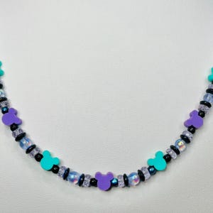 May include: A beaded necklace featuring a repeating pattern of purple and teal Mickey Mouse-shaped charms. The necklace also includes black and iridescent beads. The necklace is displayed against a white background.