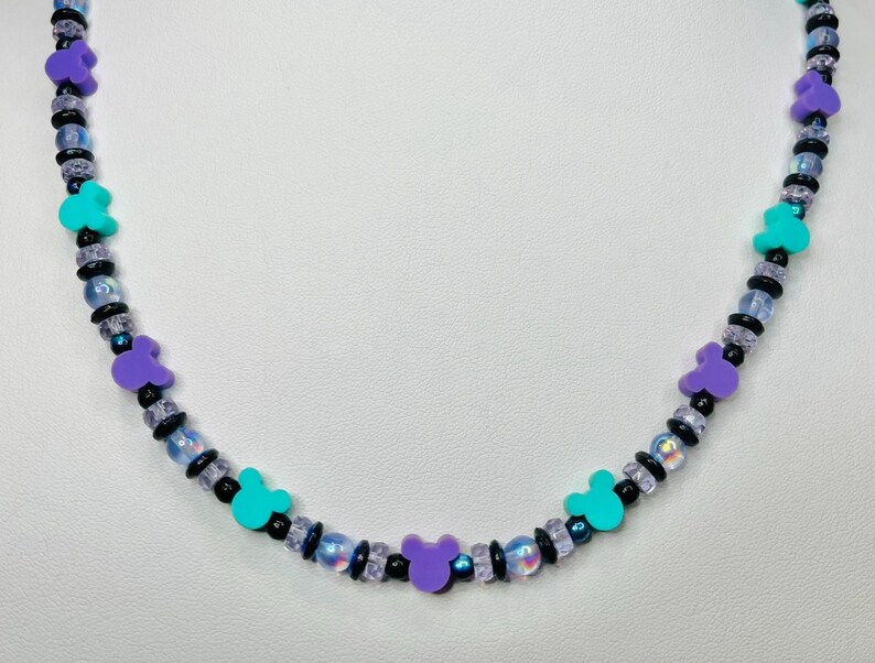 May include: A beaded necklace featuring a repeating pattern of purple and teal Mickey Mouse-shaped charms. The necklace also includes black and iridescent beads. The necklace is displayed against a white background.