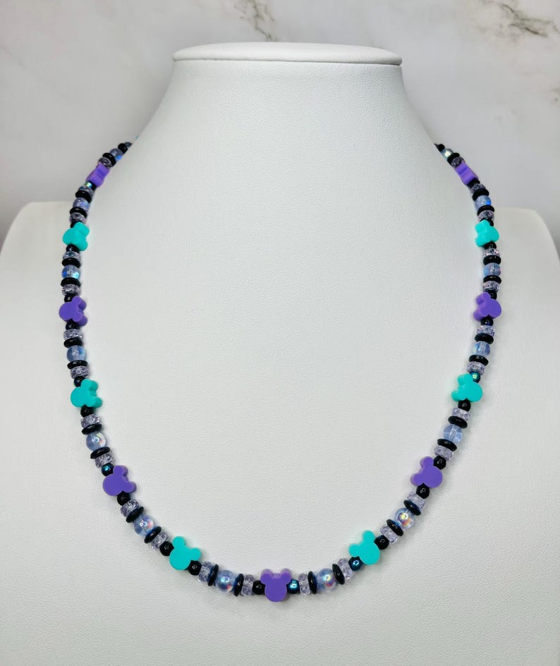 May include: A beaded necklace with a playful design. The necklace features alternating beads in black, iridescent, and small turquoise and purple Mickey Mouse-shaped beads. The necklace is displayed on a white mannequin.