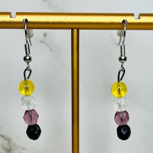 May include: A pair of dangle earrings with a non-binary pride flag design. Each earring features a yellow, white, lavender, and black bead design, suspended from a silver-colored hook. The earrings are displayed on a gold-colored earring stand.