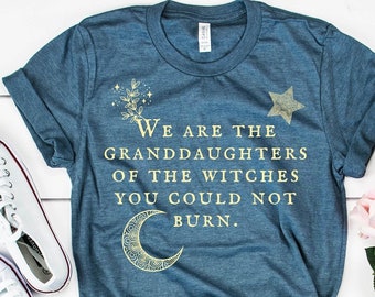 May include: A heather slate blue t-shirt with the text "We are the granddaughters of the witches you could not burn." The shirt is paired with denim shorts and white sneakers. A bouquet of pink and white roses is also present.