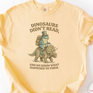 Peut inclure: T-shirt jaune beurre avec le texte "DINOSAURS DIDN'T READ, AND WE KNOW WHAT HAPPENED TO THEM." Une grenouille en haut-de-forme monte un dinosaure. Le t-shirt a des manches courtes et un col rond.